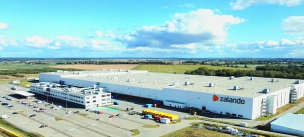 Zalando warehouse facility with blue and white buildings in rural landscape under clear sky with clouds